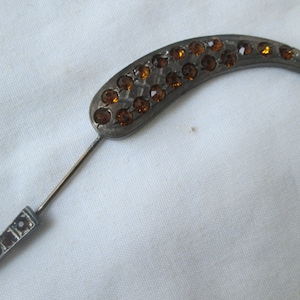 May include: A silver-toned pin with a curved, teardrop-shaped head. The head is decorated with a cluster of amber-colored rhinestones.