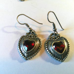 May include: A pair of silver-toned drop earrings featuring a heart-shaped design with a deep red faceted stone at the center. The metal frame has a textured, beaded border and vertical line detailing, suspended from simple silver-toned ear wires.