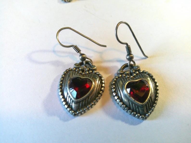 May include: A pair of silver-toned drop earrings featuring a heart-shaped design with a deep red faceted stone at the center. The metal frame has a textured, beaded border and vertical line detailing, suspended from simple silver-toned ear wires.