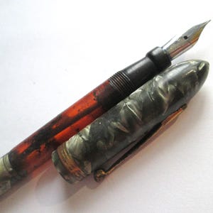 May include: An antique fountain pen with a black and marbled gray barrel, a dark red-brown body, and a silver nib. The pen's cap is open, revealing the nib and the ink reservoir. The pen has a gold-colored clip.