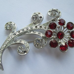 May include: A silver brooch with a red rhinestone flower design. The flower has a cluster of red rhinestones in the center and is surrounded by a silver filigree design.