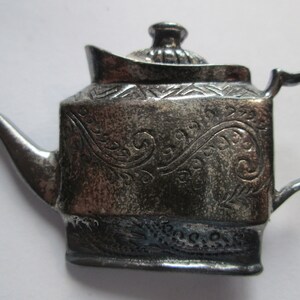 May include: A silver teapot with an ornate design. The teapot has a lid with a knob and a spout. The teapot is decorated with a floral pattern.