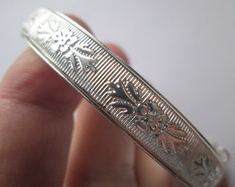 May include: Silver bangle bracelet with a decorative floral pattern. The bracelet is smooth and shiny.