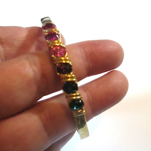 May include: A gold-toned bracelet with a row of colorful gemstones. The gemstones are in shades of red, pink, purple, green, and clear. The bracelet is held in a hand against a white background. The gemstones are set in a gold wire design.