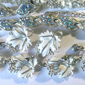 May include: A collection of vintage jewelry including two silver-tone bracelets and matching earrings. One bracelet features iridescent blue rhinestones in leaf-shaped settings, while the other bracelet and earrings display white enamel grape leaves.