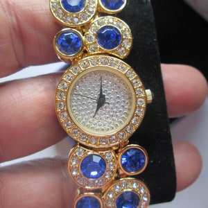 May include: A gold-tone watch with a white face and blue crystal accents. The watch has a round face and a band made of linked circles with blue crystals.