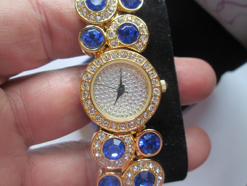 May include: A gold-tone watch with a white face and blue crystal accents. The watch has a round face and a band made of linked circles with blue crystals.