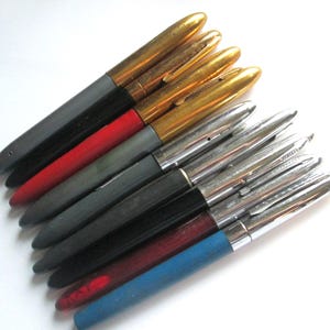 May include: A collection of vintage fountain pens with gold, silver, black, red, and blue barrels. The pens have gold or silver-colored nibs and caps, arranged diagonally against a white background.
