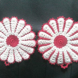 May include: Two handmade crochet flower-shaped potholders. Each potholder features a red center and a white petal design with red edging. The potholders are displayed on a dark surface.
