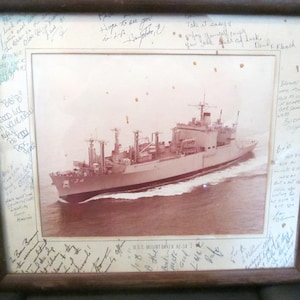 May include: Framed sepia-toned photograph of the U.S.S. Mount Baker, a naval ship, sailing on the water. The image is surrounded by a dark wooden frame and handwritten signatures and messages.