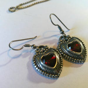 May include: A pair of silver-toned drop earrings featuring a heart-shaped design with a deep red faceted stone at the center. The metal frame has a textured, beaded border and a dark, oxidized finish. Each earring hangs from a simple silver hook wire.