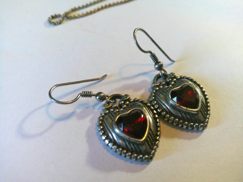 May include: A pair of silver-toned drop earrings featuring a heart-shaped design with a deep red faceted stone at the center. The metal frame has a textured, beaded border and a dark, oxidized finish. Each earring hangs from a simple silver hook wire.