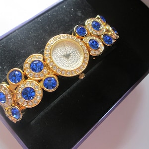 May include: A gold-tone watch with a white face and blue crystal accents. The watch has a round face and a bracelet-style band.