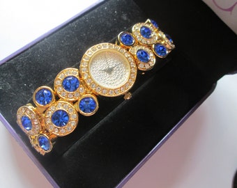 May include: A gold-tone watch with a white face and blue crystal accents. The watch has a round face and a bracelet-style band.