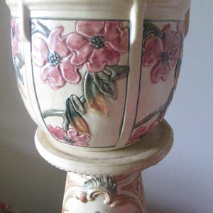 May include: A vintage ceramic planter with a floral design. The planter is white with pink and green flowers and leaves. It is sitting on a matching pedestal.
