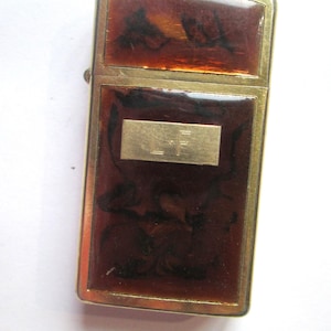 May include: Vintage lighter with a rectangular shape. The lighter features a gold-colored metal frame and panels with a brown, marbled design. A gold-colored rectangular plate with the initials "L.F." is centered on the front.