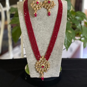 May include: A red beaded necklace with a gold pendant and matching earrings. The pendant and earrings feature a floral design with pink stones.