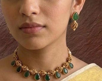 Könnte beinhalten: Ein goldfarbenes Choker-Halsband und passende Ohrhänger mit tropfenförmigen grünen Steinen und Clustern aus kleinen Goldperlen. Das Schmuckset wird auf einem cremefarbenen Stoff mit Goldrand getragen und enthält den Schriftzug Era.