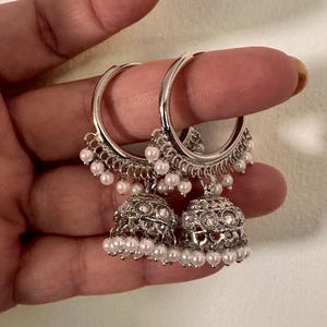 May include: Silver-toned hoop earrings with dangling bell-shaped charms. The charms are adorned with small white pearls and intricate detailing. The earrings are designed with a delicate and elegant aesthetic, suitable for formal or casual wear.