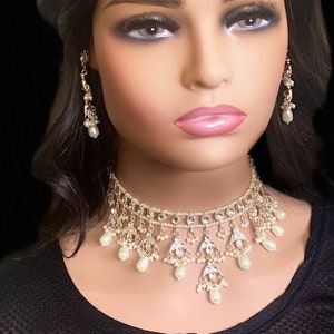 May include: A white and gold bridal jewellery set with pearl accents. The set includes a choker necklace, earrings, and a tikka.