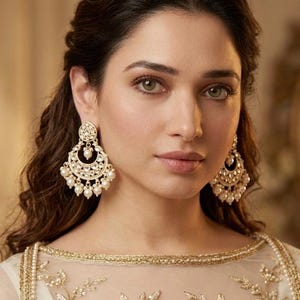 May include: A woman wearing gold-colored chandelier earrings with pearl accents. The earrings are ornate and detailed. She is wearing a white and gold embroidered top. The image is a close-up portrait.