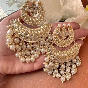 May include: Gold-toned, ornate earrings with a crescent shape, adorned with small, clear stones and dangling faux pearls. The earrings have a layered design and are held in a hand, showcasing their intricate details.