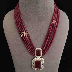 May include: A necklace and earring set featuring ruby red beads and stones. The necklace has multiple strands of small, round beads, with a large rectangular pendant and a smaller stone above it. The earrings mirror the rectangular design. The set is displayed on a mannequin.