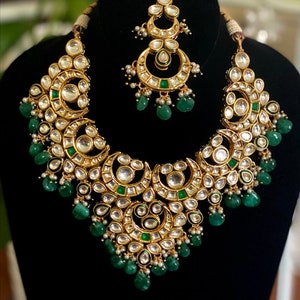 May include: A gold-toned necklace and headpiece set. The necklace features a crescent moon design with clear and green accents, and dangling green beads. The headpiece has a similar design with dangling pearls.