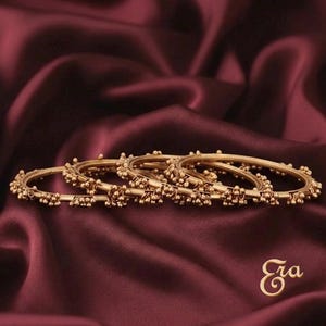 May include: A set of three gold-toned metal bangles, each featuring a thin circular band adorned with small, clustered spherical beads spaced evenly around the circumference. The jewellery is arranged in a row, displaying a metallic finish.