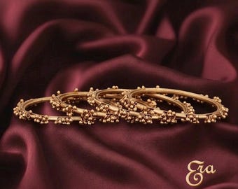 May include: A set of three gold-toned metal bangles, each featuring a thin circular band adorned with small, clustered spherical beads spaced evenly around the circumference. The jewelry is arranged in a row, displaying a metallic finish.