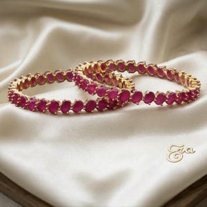 May include: Two circular bangles featuring a continuous row of oval-shaped, deep pink stones set in a gold-toned metal frame. The jewelry pieces are arranged overlapping each other. The text "Eva" is visible in the lower right corner.