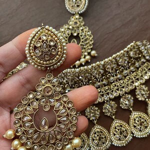 May include: A gold-tone necklace and earrings set with clear stones and pearl accents. The necklace is a choker style with a layered design and the earrings are teardrop shaped.
