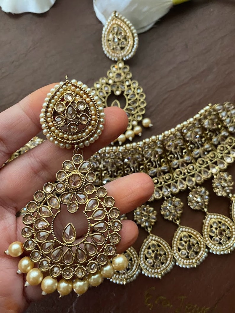 May include: A gold-tone necklace and earrings set with clear stones and pearl accents. The necklace is a choker style with a layered design and the earrings are teardrop shaped.