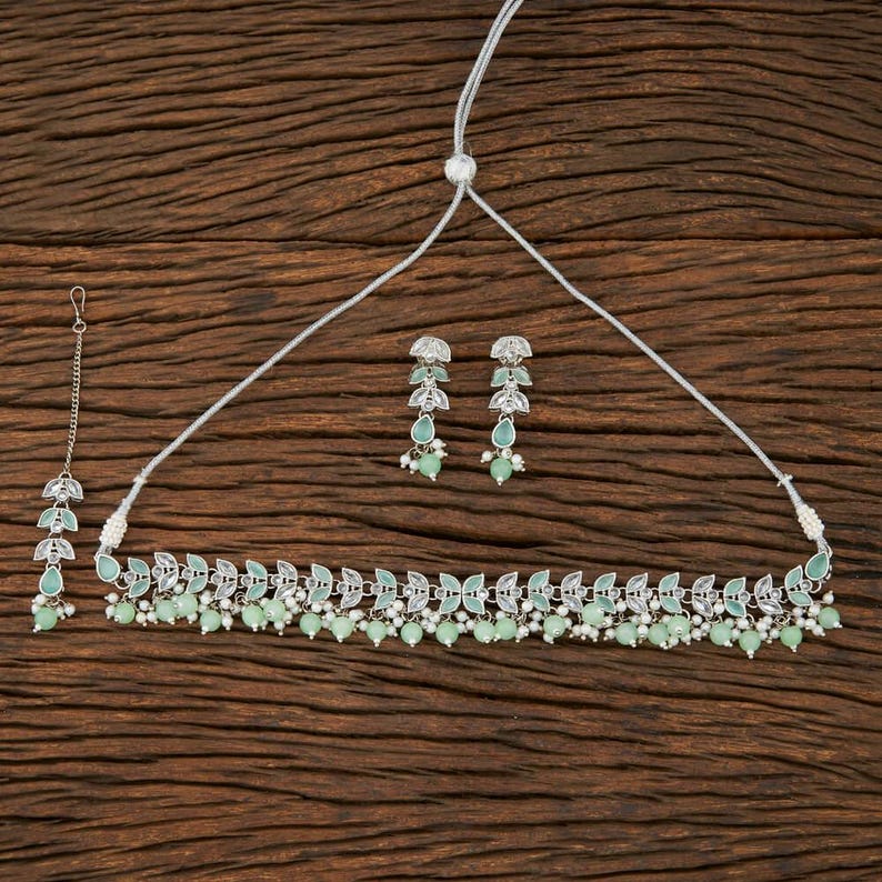 May include: A silver-toned jewelry set featuring a necklace, earrings, and a headpiece. The necklace and earrings have a leaf design with light green accents and small, round beads. The headpiece has a similar leaf design.