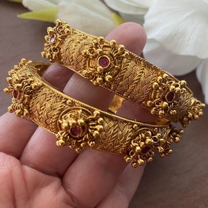 Può includere: Due braccialetti d'oro con disegni intricati e pietre preziose rosse. I braccialetti sono impilati uno sopra l'altro.