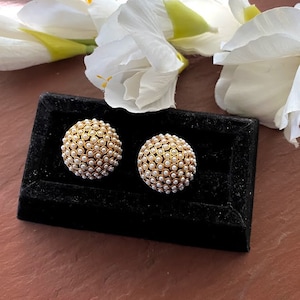 May include: A pair of gold tone earrings with white pearl accents. The earrings are round and have a textured surface. The earrings are displayed on a black velvet background.