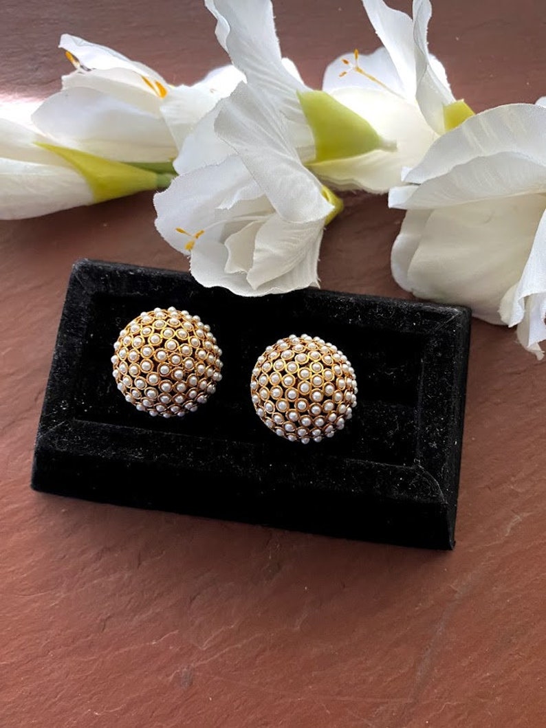 May include: A pair of gold tone earrings with white pearl accents. The earrings are round and have a textured surface. The earrings are displayed on a black velvet background.