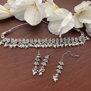 May include: A silver-toned jewelry set featuring a choker necklace, earrings, and a headpiece. The necklace and earrings have a leaf design with clear stones and small white pearls. The headpiece has a similar leaf design. White flowers are in the background.