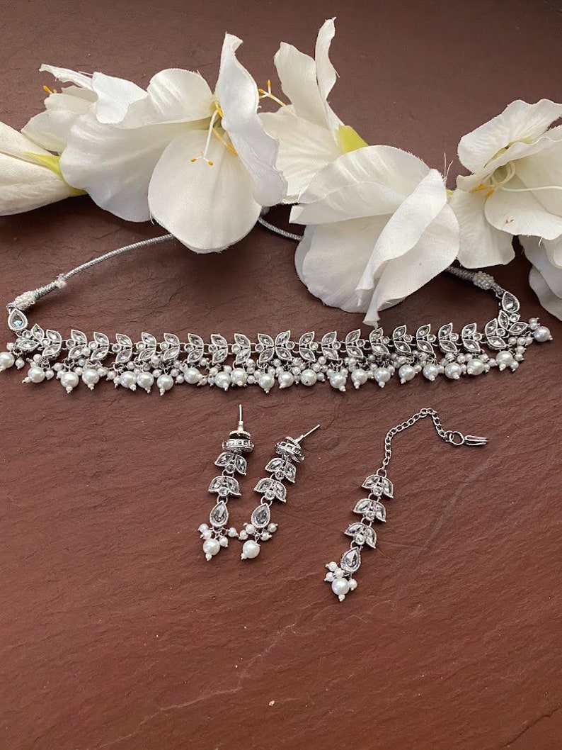 May include: A silver-toned jewelry set featuring a choker necklace, earrings, and a headpiece. The necklace and earrings have a leaf design with clear stones and small white pearls. The headpiece has a similar leaf design. White flowers are in the background.