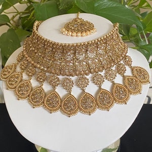May include: A gold-tone bridal necklace set with white stones and pearls. The set includes a choker necklace, a matching maang tikka, and dangling earrings.