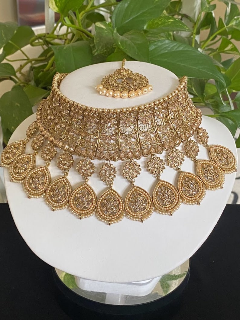 May include: A gold-tone bridal necklace set with white stones and pearls. The set includes a choker necklace, a matching maang tikka, and dangling earrings.