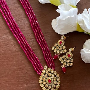 May include: A pink beaded necklace with a gold pendant and matching earrings. The pendant is adorned with clear and red stones.