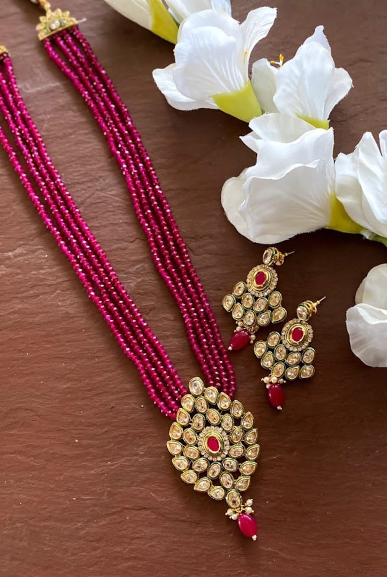 May include: A pink beaded necklace with a gold pendant and matching earrings. The pendant is adorned with clear and red stones.