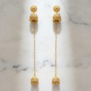 May include: A pair of gold-colored dangle earrings. Each earring features a circular top with a central gem, a textured bell-shaped body, and a long chain ending in a smaller bell. The earrings are displayed against a white marble background.