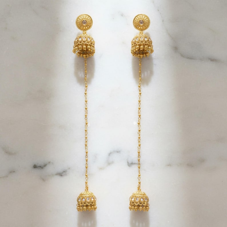 May include: A pair of gold-colored dangle earrings. Each earring features a circular top with a central gem, a textured bell-shaped body, and a long chain ending in a smaller bell. The earrings are displayed against a white marble background.