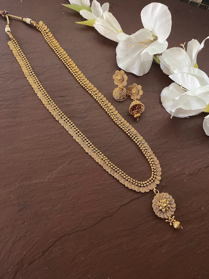 May include: Gold-toned necklace and matching earrings set. The necklace features a long chain with coin-like pendants and a detailed pendant at the bottom. The earrings have a floral design with a dangling element. The jewelry is displayed on a brown surface.