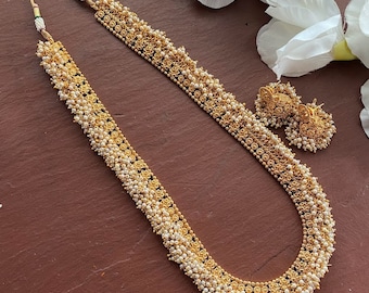 May include: A gold-toned necklace and matching earrings set. The necklace is long, with a central section of small, round, white beads and gold floral accents. The earrings have a similar design. The jewelry is displayed on a brown surface.