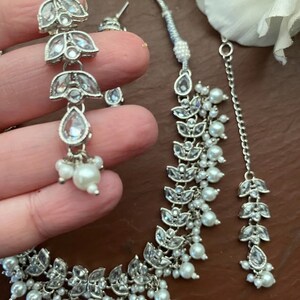 May include: Silver-toned jewelry set featuring a necklace, earrings, and a headpiece. The set includes clear, sparkling accents and small, round, white pearls. The earrings are long and dangle, while the necklace and headpiece have a floral design.