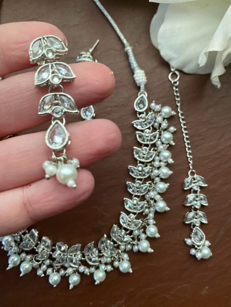 May include: Silver-toned jewelry set featuring a necklace, earrings, and a headpiece. The set includes clear, sparkling accents and small, round, white pearls. The earrings are long and dangle, while the necklace and headpiece have a floral design.