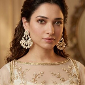 May include: A woman wearing gold-colored chandelier earrings with pearl accents. The earrings are ornate and detailed. She is wearing a white and gold embroidered top. The image is a close-up portrait.